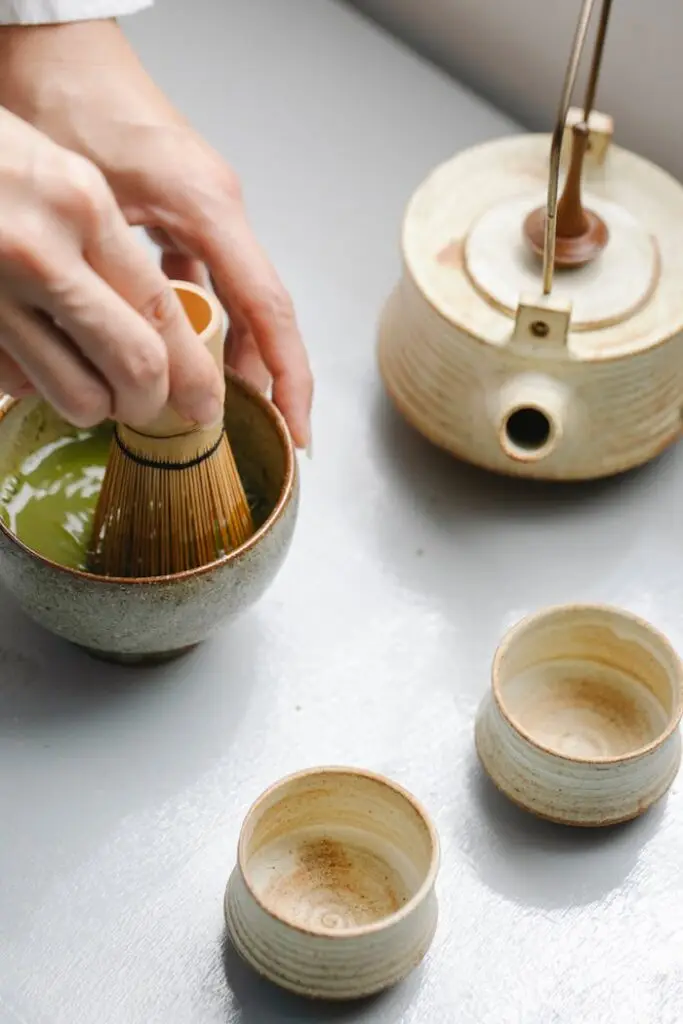 From above of crop person mixing matcha powder in bowl with bamboo whisk while brewing healthy tea on table with teapot