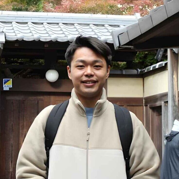 A young man, Kenjiro Takeda, smiling in front of a traditional Japanese building with autumn foliage in the background.